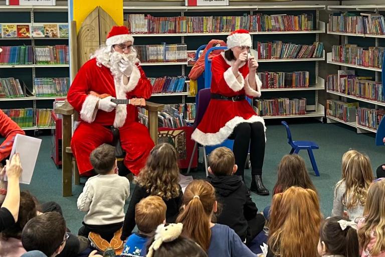 Two people dressed as Santa Claus in a library with children seated 
