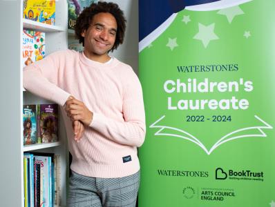 Joseph Coelho leaning on a bookshelf with a banner to his right saying he is Waterstones Children's Laureate 2023 to 2024