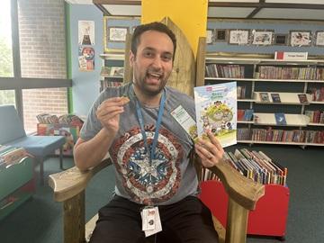 A Library Assistant with Summer Reading Materials including a medal, sitting in a chair in a library
