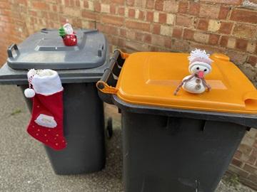 An orange-lidded bin with a toy snowman on top, and a black-lidded bin with a toy Father Christmas on top and a stocking hanging from the lid