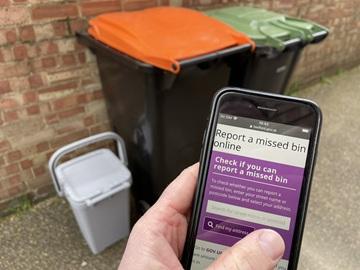 A mobile phone showing the missed bin webpage, with bins in the background