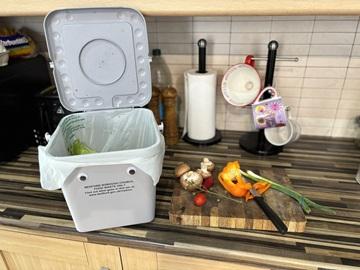 A kitchen food waste caddy, with googly eyes, on a kitchen side, next to to a chopping board with veg