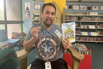 A Library Assistant with Summer Reading Materials including a medal, sitting in a chair in a library