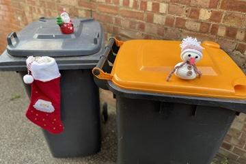An orange-lidded bin with a toy snowman on top, and a black-lidded bin with a toy Father Christmas on top and a stocking hanging from the lid