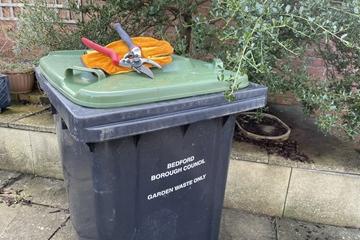 Green-lidded bin with gardening tools on lid and bush behind