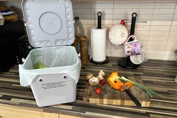 A kitchen food waste caddy, with googly eyes, on a kitchen side, next to to a chopping board with veg